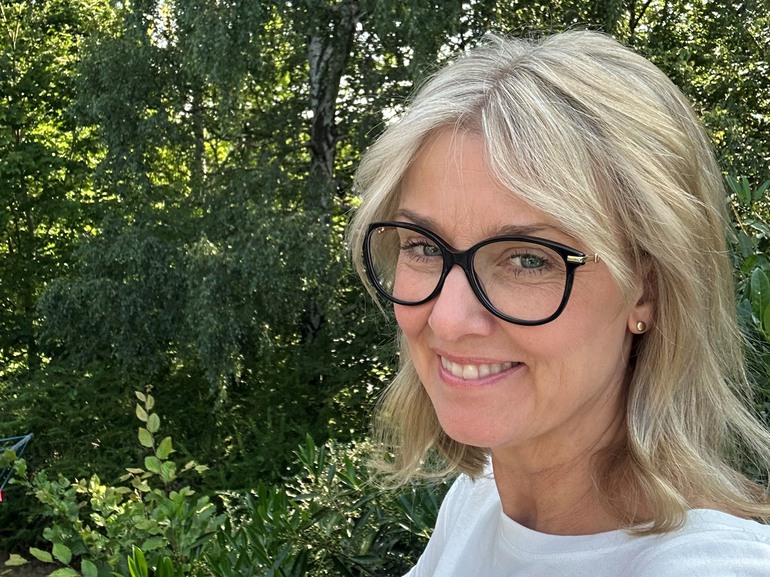 kvinna med glasögon och ljust halvlångt hår tittar in i kameran i en grönskande miljö