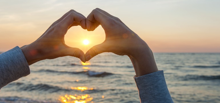 Två händer som formar ett hjärta framför en solnedgång över havet.