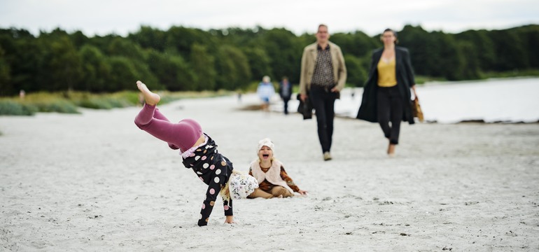 Två barn som leker vid en strand. I bakgrunden syns en man och en kvinna som går mot barnen.