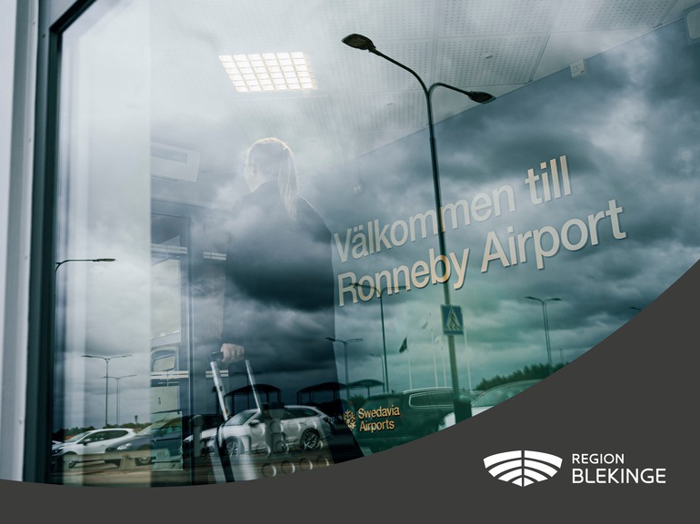 Bild på en person som går med resväska , framför en en vägg där det står "Välkommen till Ronneby Airport". 