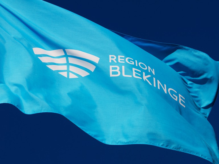 En närbild på en blå flagga med Region Blekinges logotyp mot en klarblå himmel.