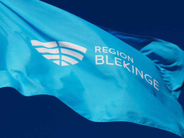 En närbild på en blå flagga med Region Blekinges logotyp mot en klarblå himmel.