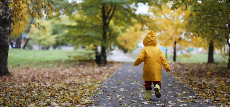 Ett barn i gul höstjacka går på asfalt. I bakgrunden syns träd och gula höstlöv på marken. 
