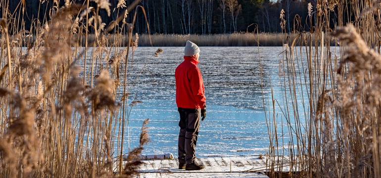 En man i röd jacka, svarta byxor och skor, och en grå mössa, står framför en sjö. Runtom syns vass. 