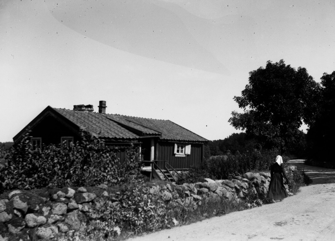 Äldre svartvitt foto föreställande låg stuga med stenmur med en dam med huvudduk gående bakifrån