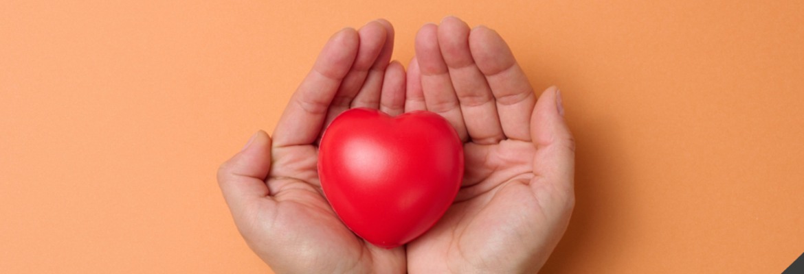 Bild på två händer mot orange bakgrund, som håller i ett rött hjärta av plast. 