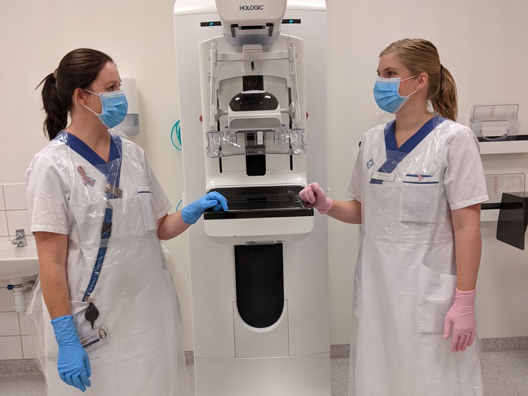 Två personer ur personalen står vid en röntgenapparat som används för mammografi. De har på sig munskydd, plastförkläde och handskar.