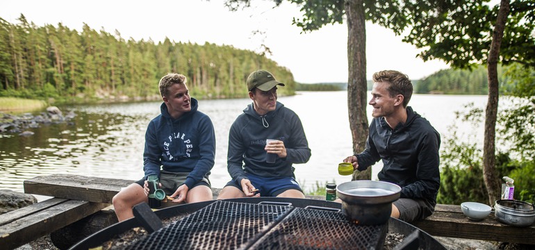 Tre personer sitter på en bänk framför en eldstad och lagar maten över elden. I bakgrunden syns vatten och skog.