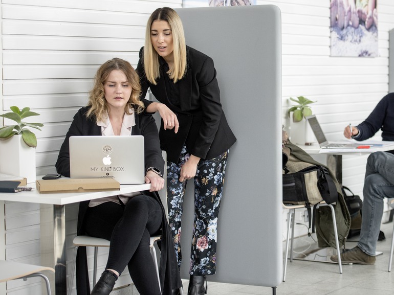 En kvinna sitter och en kvinna står vid laptop i kontorsmiljö